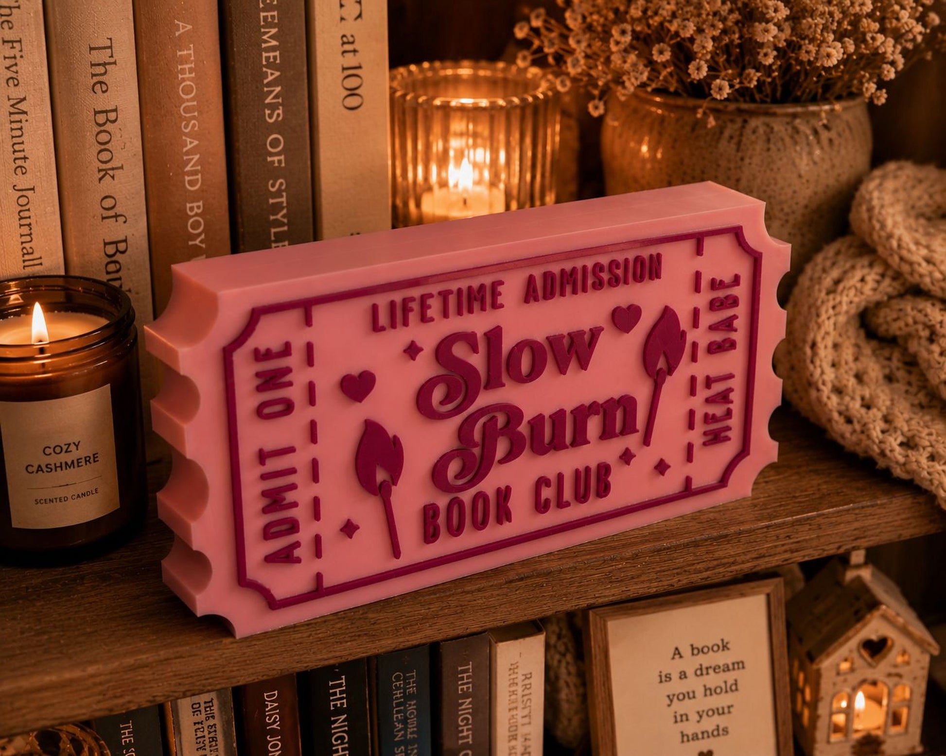 a pink ticket-shaped object with the text "LIFETIME ADMISSION Slow Burn Book Club" on it, placed on a bookshelf among various books and decorative items.
