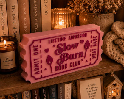 a pink ticket-shaped object with the text "LIFETIME ADMISSION Slow Burn Book Club" on it, placed on a bookshelf among various books and decorative items.