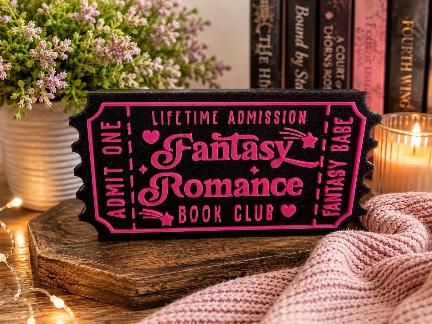 A pink and black ticket-shaped sign for a fantasy romance book club, surrounded by a potted plant, a candle, and a knitted blanket.