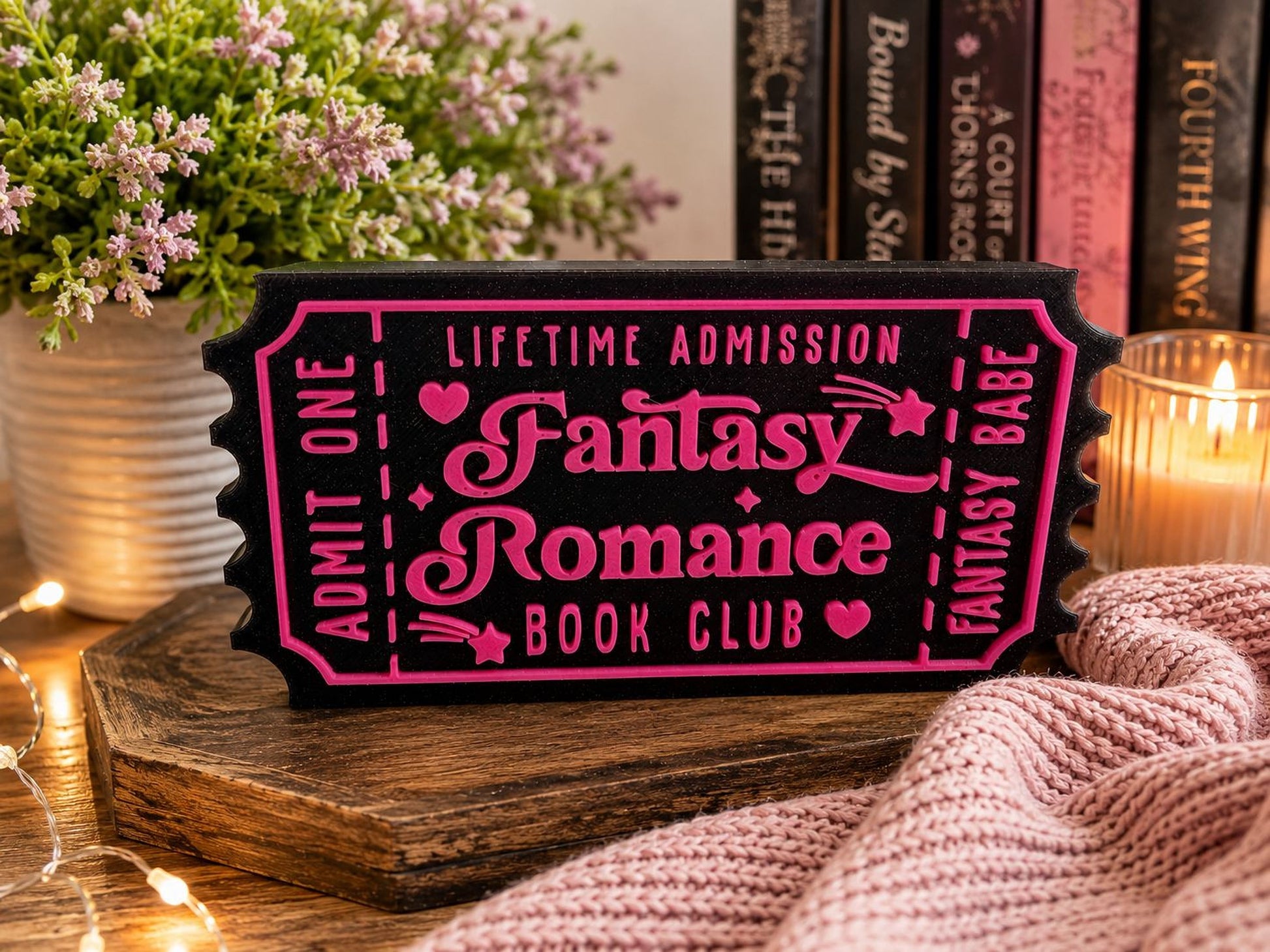A pink and black ticket-shaped sign for a fantasy romance book club, surrounded by a potted plant, a candle, and a knitted blanket.