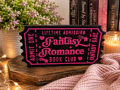A pink and black ticket-shaped sign for a fantasy romance book club, surrounded by a potted plant, a candle, and a knitted blanket.