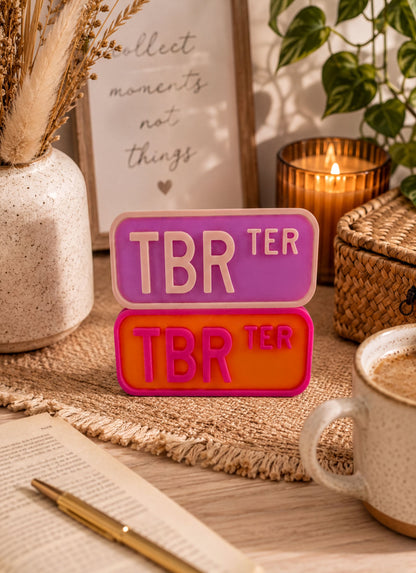 a desk with various items, including a framed quote, a mug, a pen, and a plant. The desk also has a sign that says "TBR TER" in pink and orange colors.