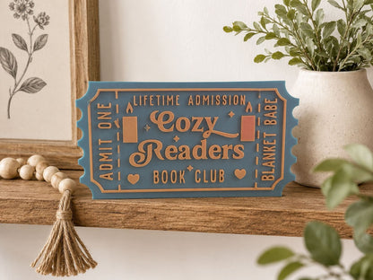 A blue ticket-shaped sign on a wooden shelf, with a tassel hanging from it, and a plant in a white vase to the right.