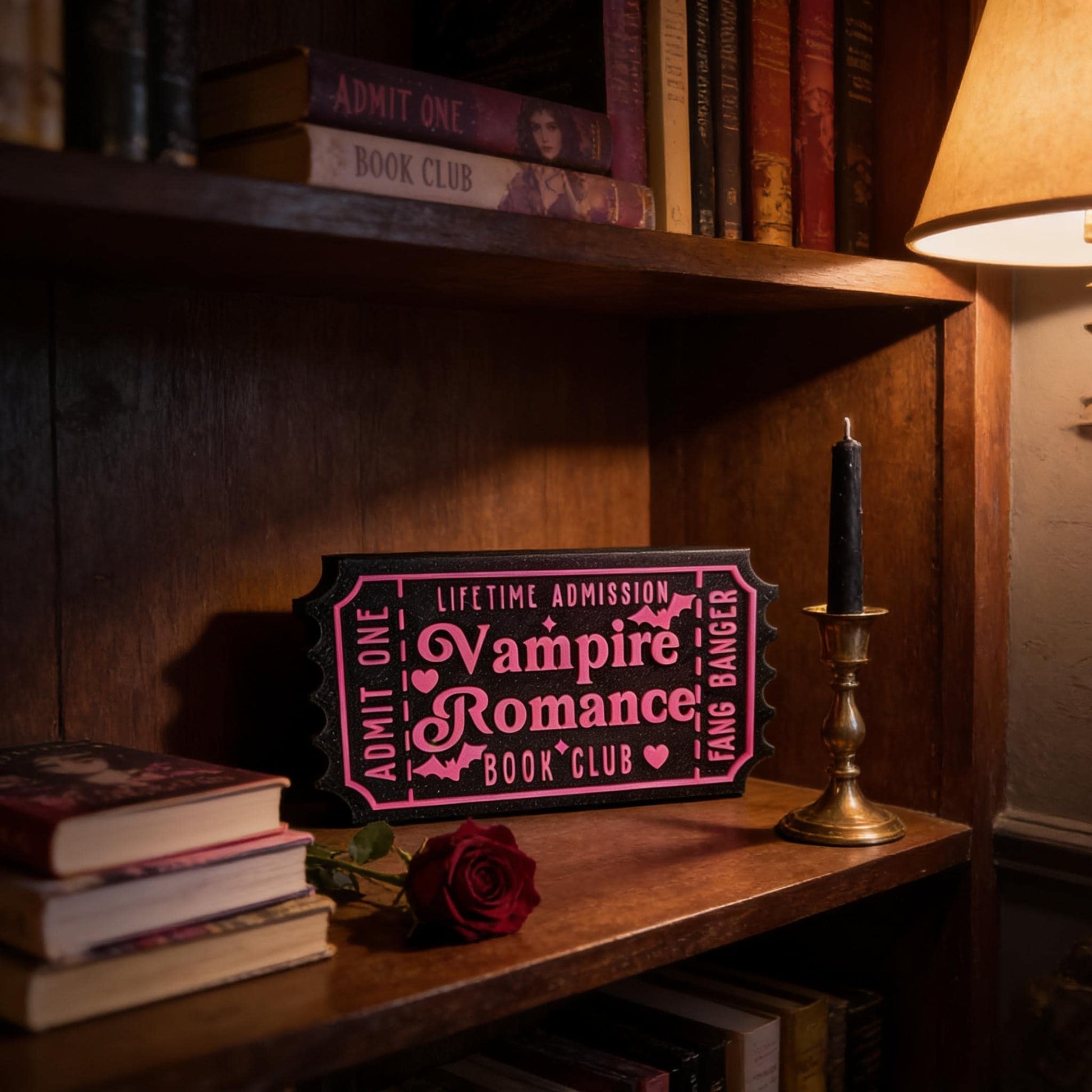 a wooden bookshelf with a pink ticket-shaped sign that reads "Lifetime Admission Vampire Romance Book Club" and a candle on the shelf.