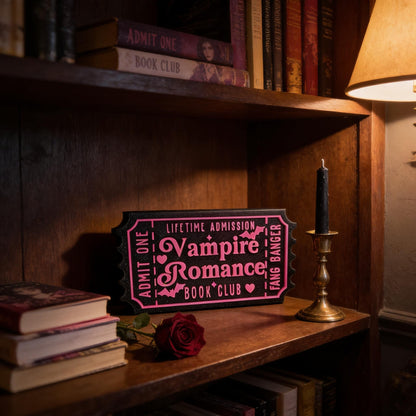 a wooden bookshelf with a pink ticket-shaped sign that reads "Lifetime Admission Vampire Romance Book Club" and a candle on the shelf.