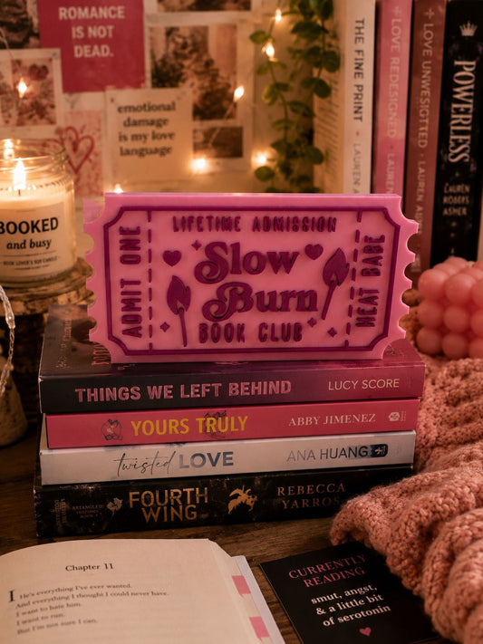 a stack of books, a pink ticket-shaped object, and some candles on a table.