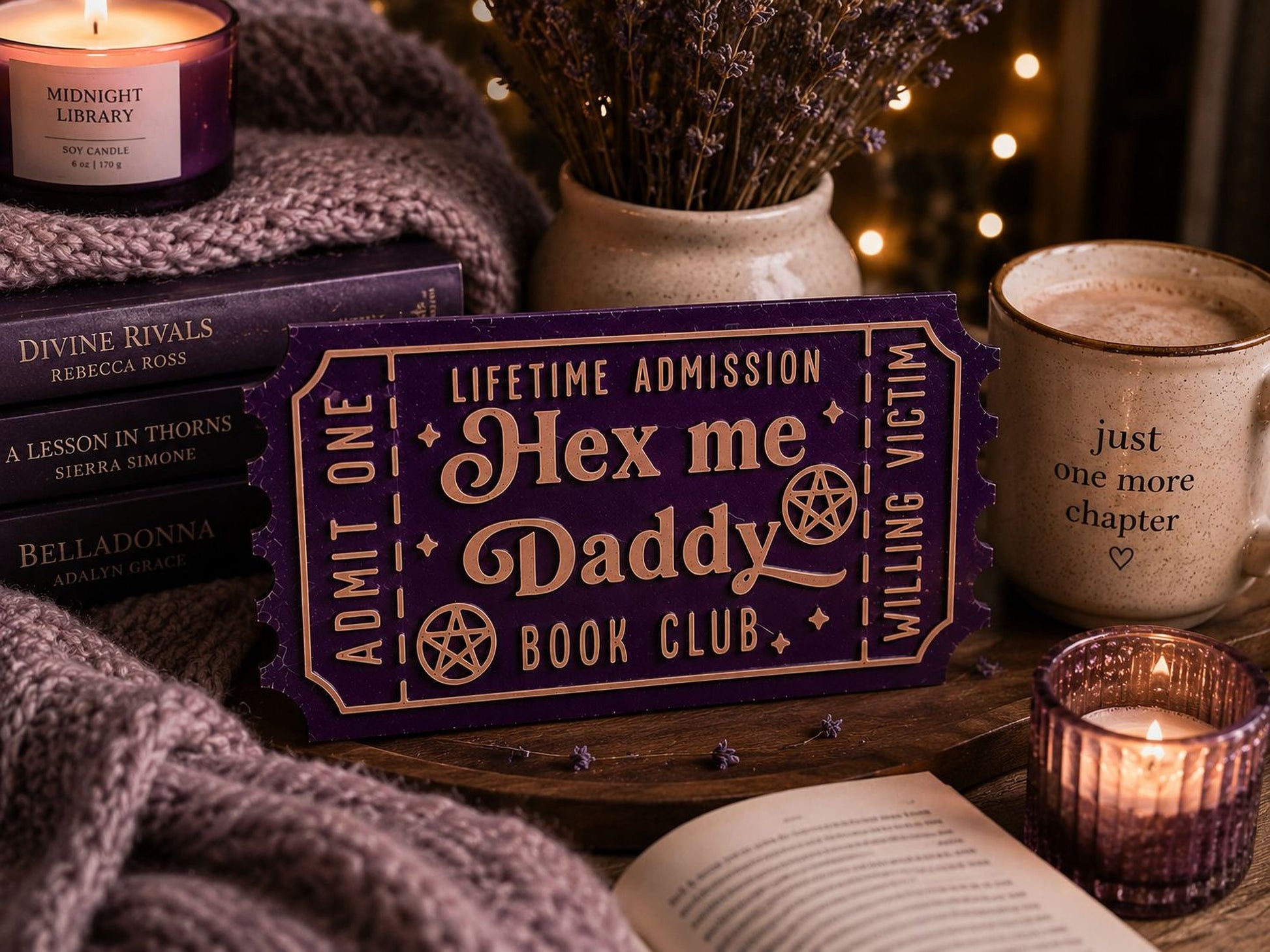 a cozy and inviting scene with a purple ticket-shaped sign that reads " Hex me Daddy Book Club" placed on a table. The table also has a stack of books, a mug, and a candle, creating a warm and comfortable atmosphere.