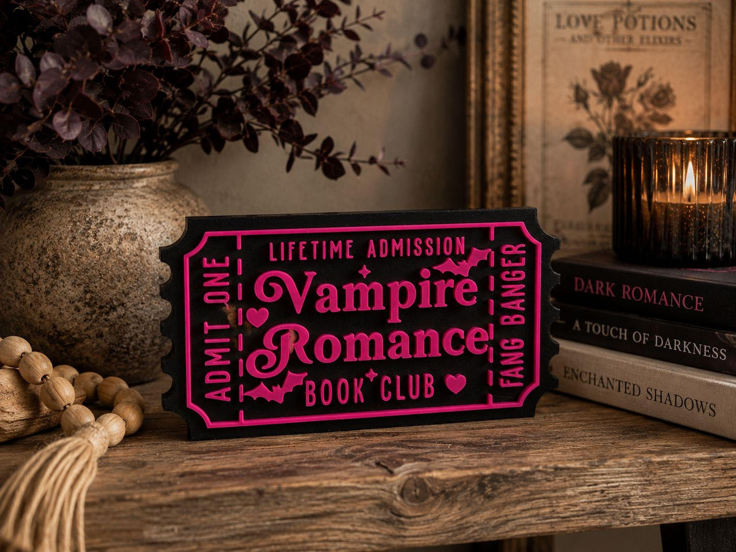a black and pink ticket-shaped sign with the words "Lifetime Admission Vampire Romance Book Club" written on it, placed on a wooden surface next to a stack of books and a candle.