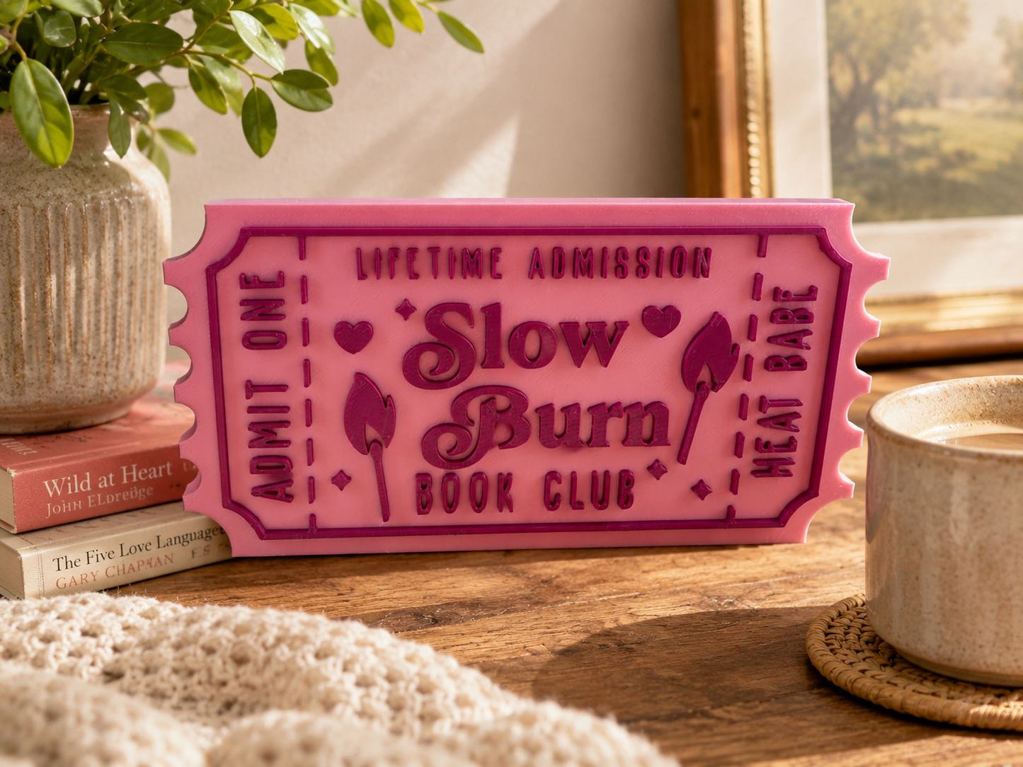 a pink ticket-shaped sign with the text "Lifetime Admission Slow Burn Book Club" on it, placed on a wooden surface next to a mug and a stack of books.