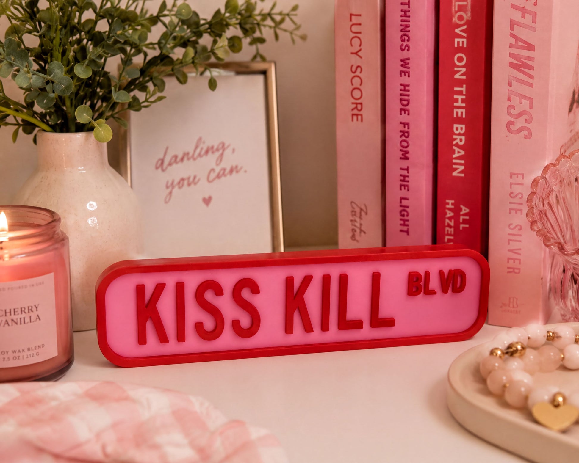 a pink and red sign that says "KISS KILL BLVD", surrounded by various pink and red items such as a candle, a framed quote, and a bracelet.