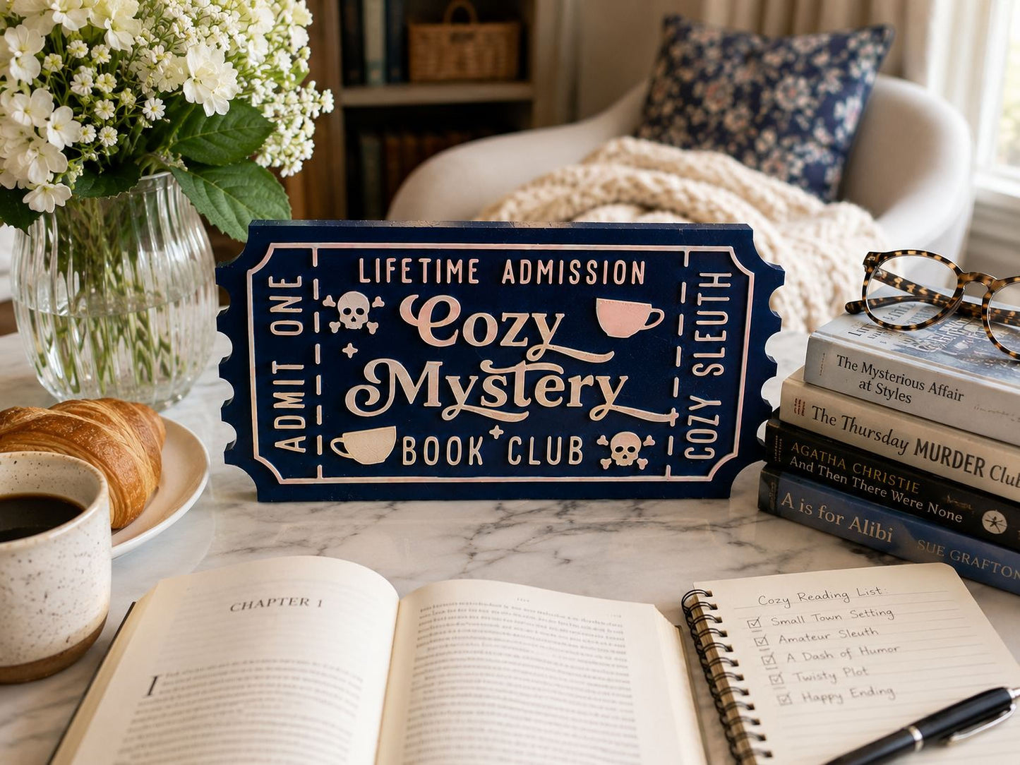 a cozy reading setup with a book club sign, a notebook, a pen, a cup of coffee, and a stack of books on a table.