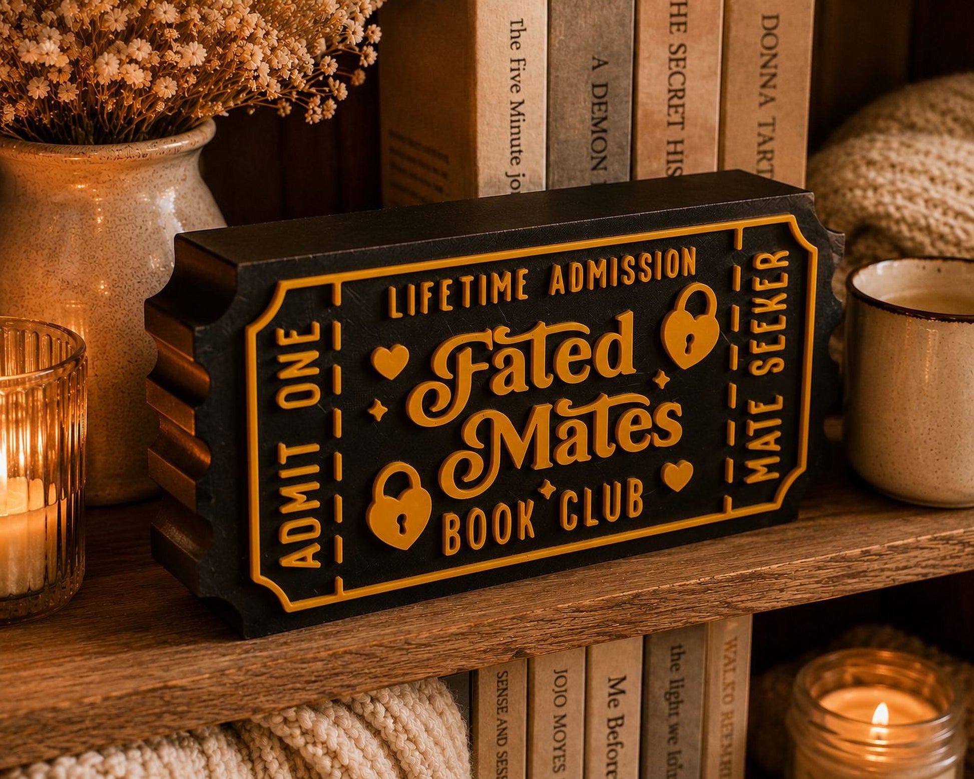 a black and gold ticket-shaped object with the text "Lifetime Admission Fated Mates Book Club" on it, placed on a wooden shelf among various books and decorative items.