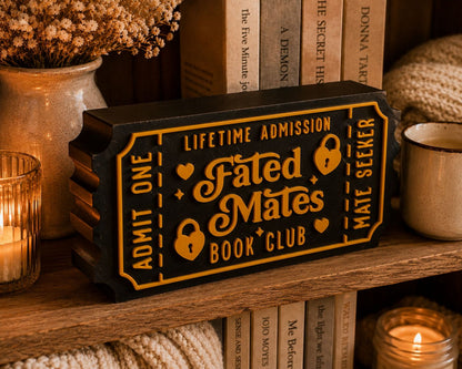 a black and gold ticket-shaped object with the text "Lifetime Admission Fated Mates Book Club" on it, placed on a wooden shelf among various books and decorative items.