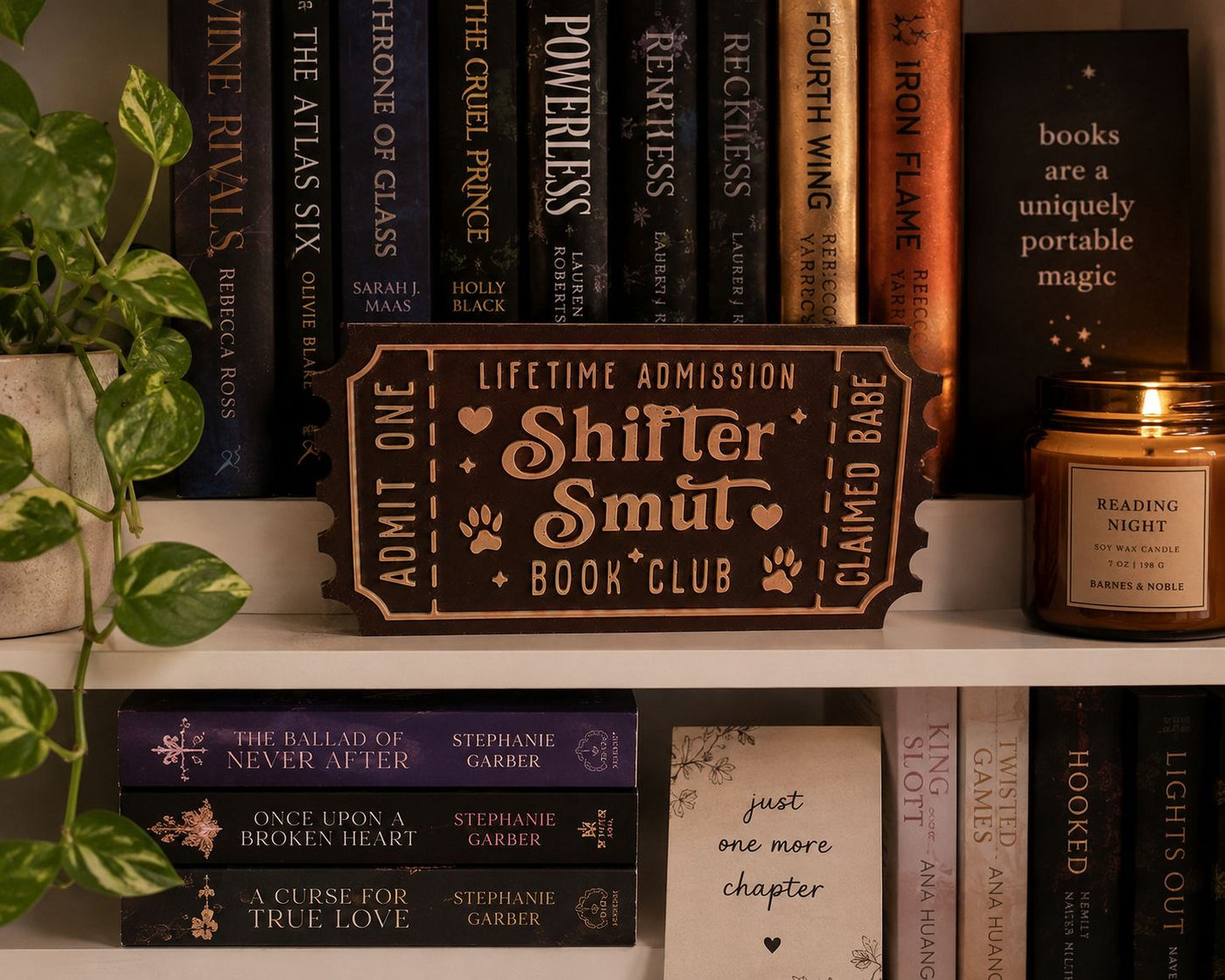 a bookshelf filled with various books, a candle, and a sign that reads "Shifter Smut Book Club".