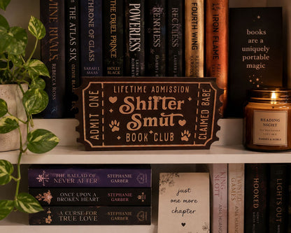 a bookshelf filled with various books, a candle, and a sign that reads "Shifter Smut Book Club".