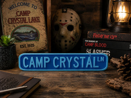 a sign that reads "CAMP CRYSTAL" in blue letters, surrounded by various items such as a mask, books, and a lantern.
