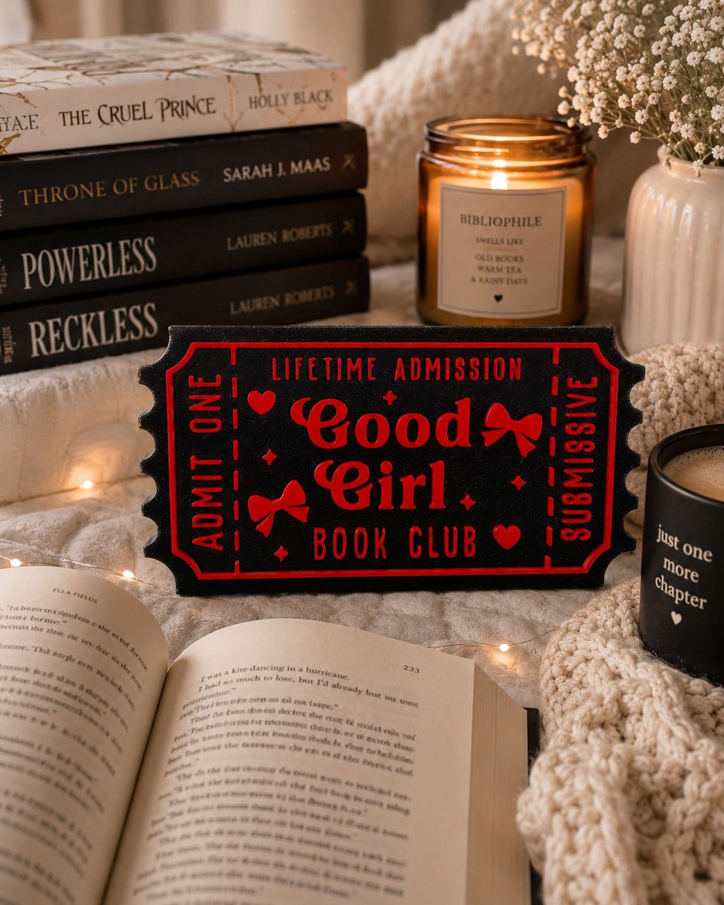 a cozy scene with a stack of books, a candle, and a book ticket sign, all arranged on a bed with a white blanket.