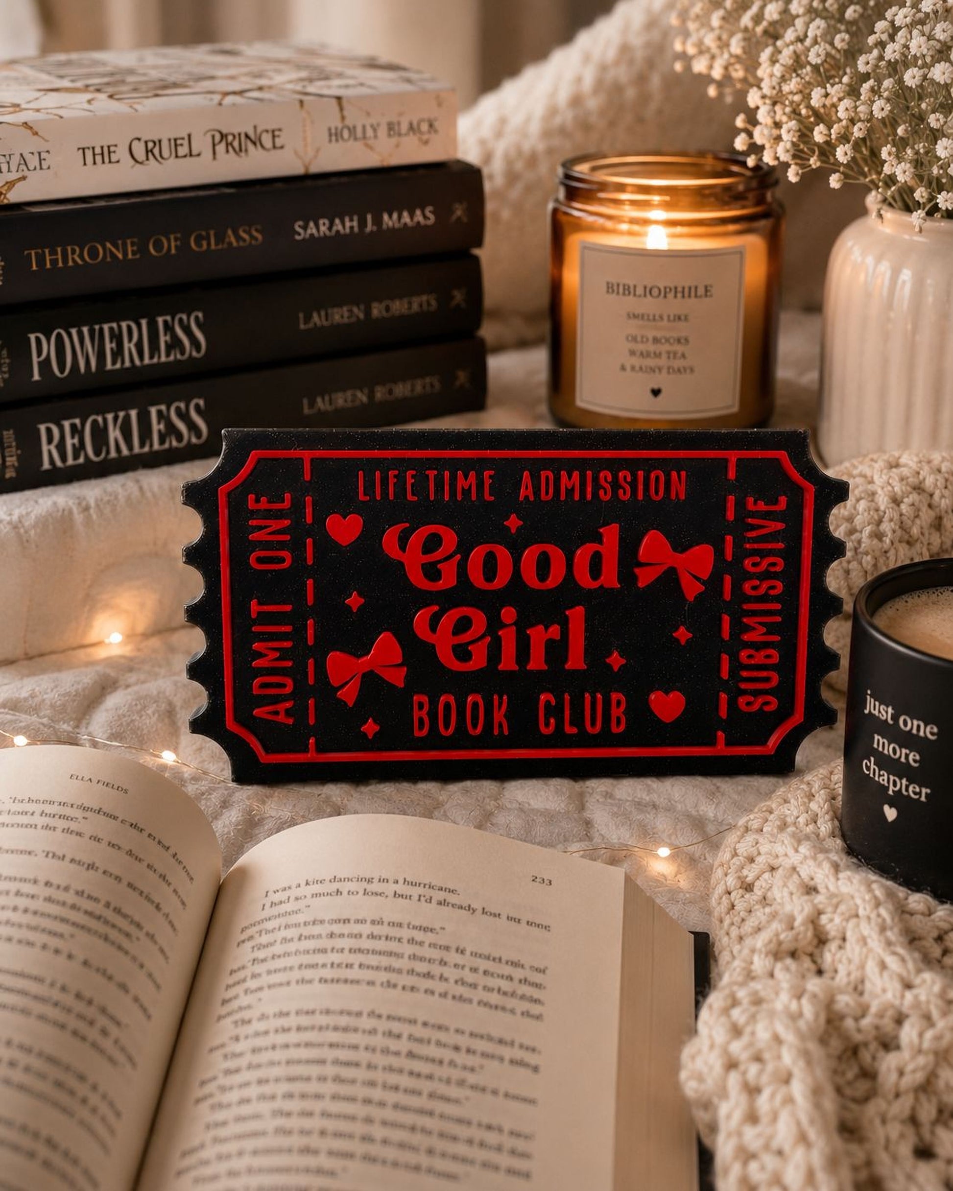 a cozy scene with a stack of books, a candle, and a book ticket sign, all arranged on a bed with a white blanket.
