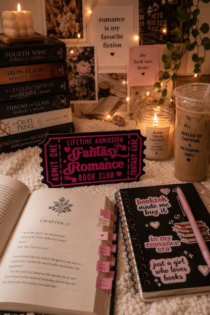 a cozy scene with a stack of romance novels, a pink ticket-shaped bookmark, a notebook, and a candle, all arranged on a bed with a white blanket.