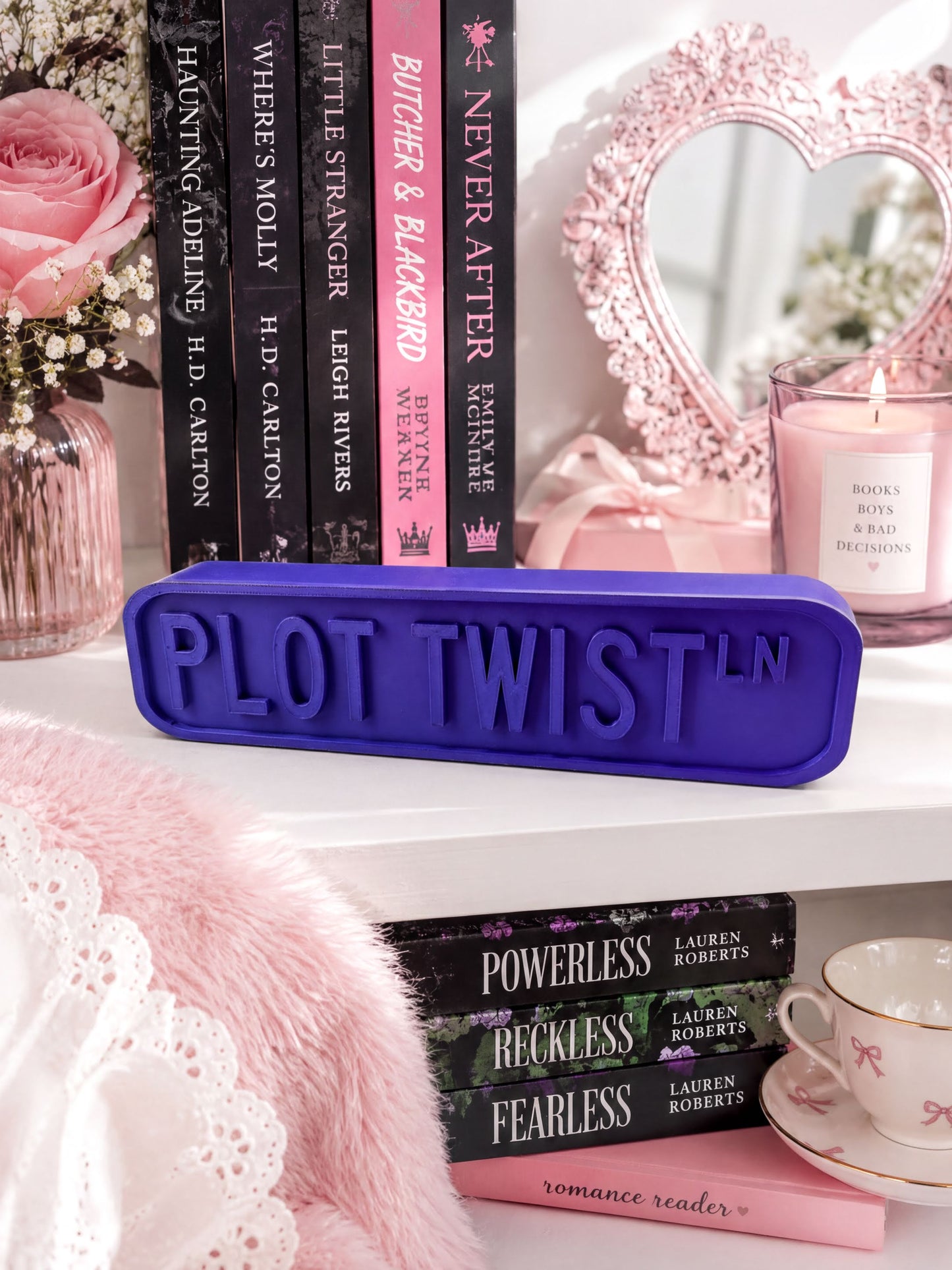 a shelf with various books, a pink candle, and a pink heart-shaped frame. The books are arranged in a row, and the shelf has a purple sign that says "PLOT TWIST".