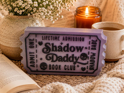a purple ticket-shaped object with the text "Lifetime Admission Shadow Daddy Book Club" on it, placed on a woven blanket next to a candle and a mug.
