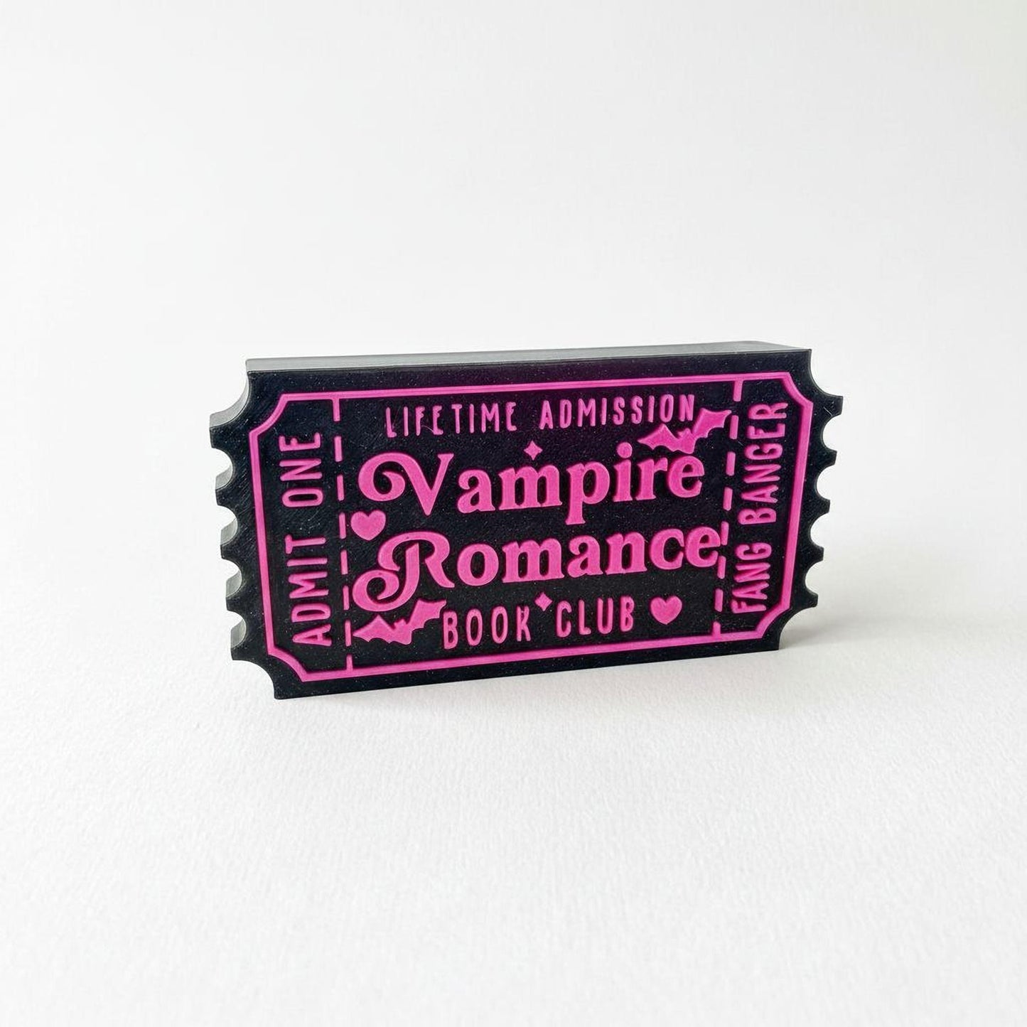 A black and pink ticket-shaped object with text that reads "Lifetime Admission Vampire Romance Book Club" printed on it.