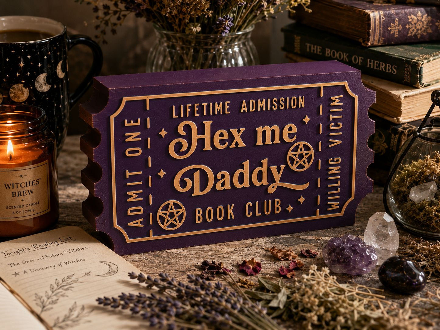 a purple ticket-shaped object with text that reads "Hex me Daddy Book Club" and "Lifetime Admission" on it, surrounded by various items such as a candle, crystals, and books.