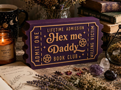a purple ticket-shaped object with text that reads "Hex me Daddy Book Club" and "Lifetime Admission" on it, surrounded by various items such as a candle, crystals, and books.