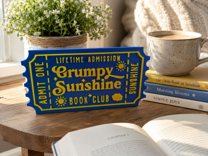 a blue ticket-shaped sign with yellow text, placed on a wooden table next to a stack of books and a mug.