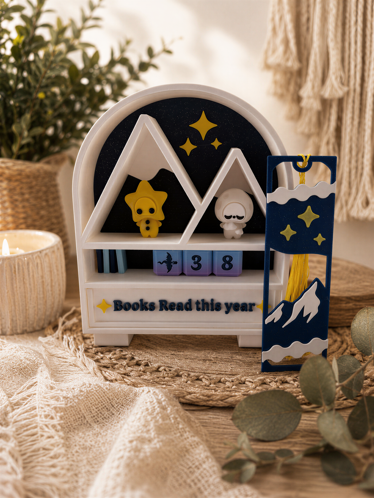 A white shelf with a blue and yellow design, displaying a small figurine and a book, surrounded by greenery and a candle.