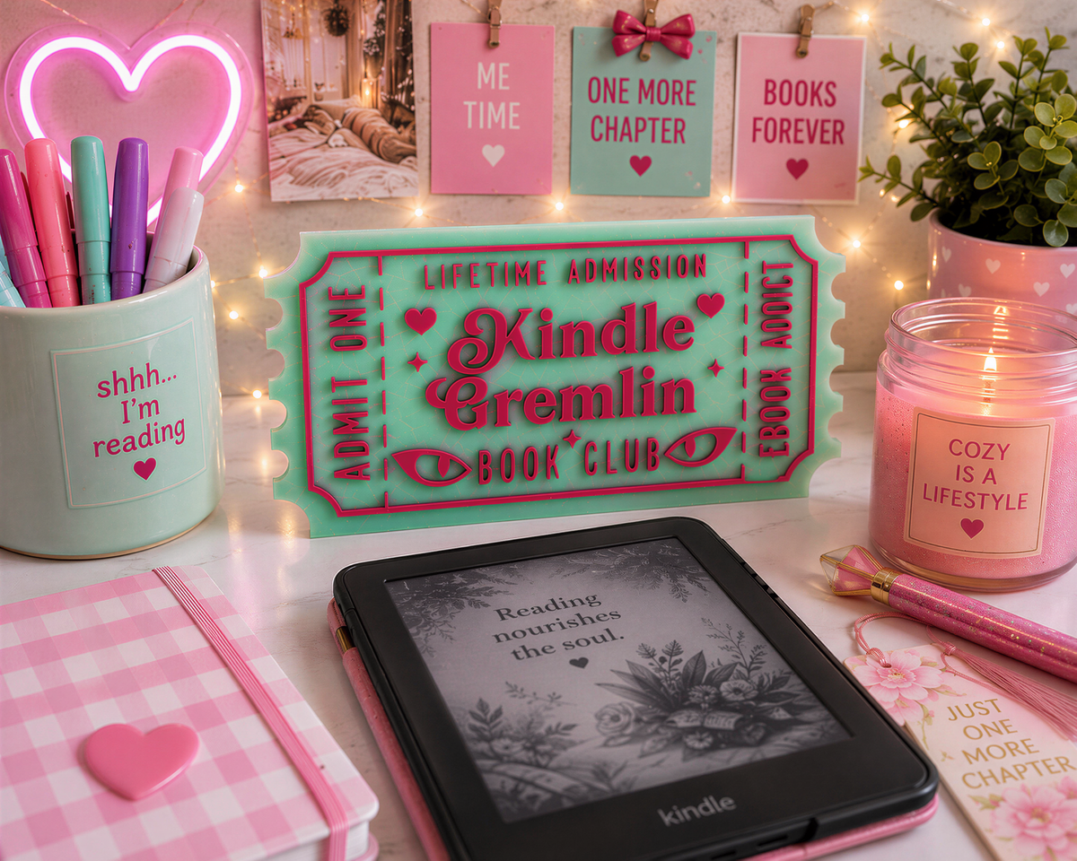 a pink and green desk with various items, including a Kindle e-reader, pens, a candle, and a heart-shaped container filled with pens. The desk also has a sign that says "KINDLE CREMLIN BOOK CLUB" and a pink candle with the text "COZY IS A LIFESTYLE".