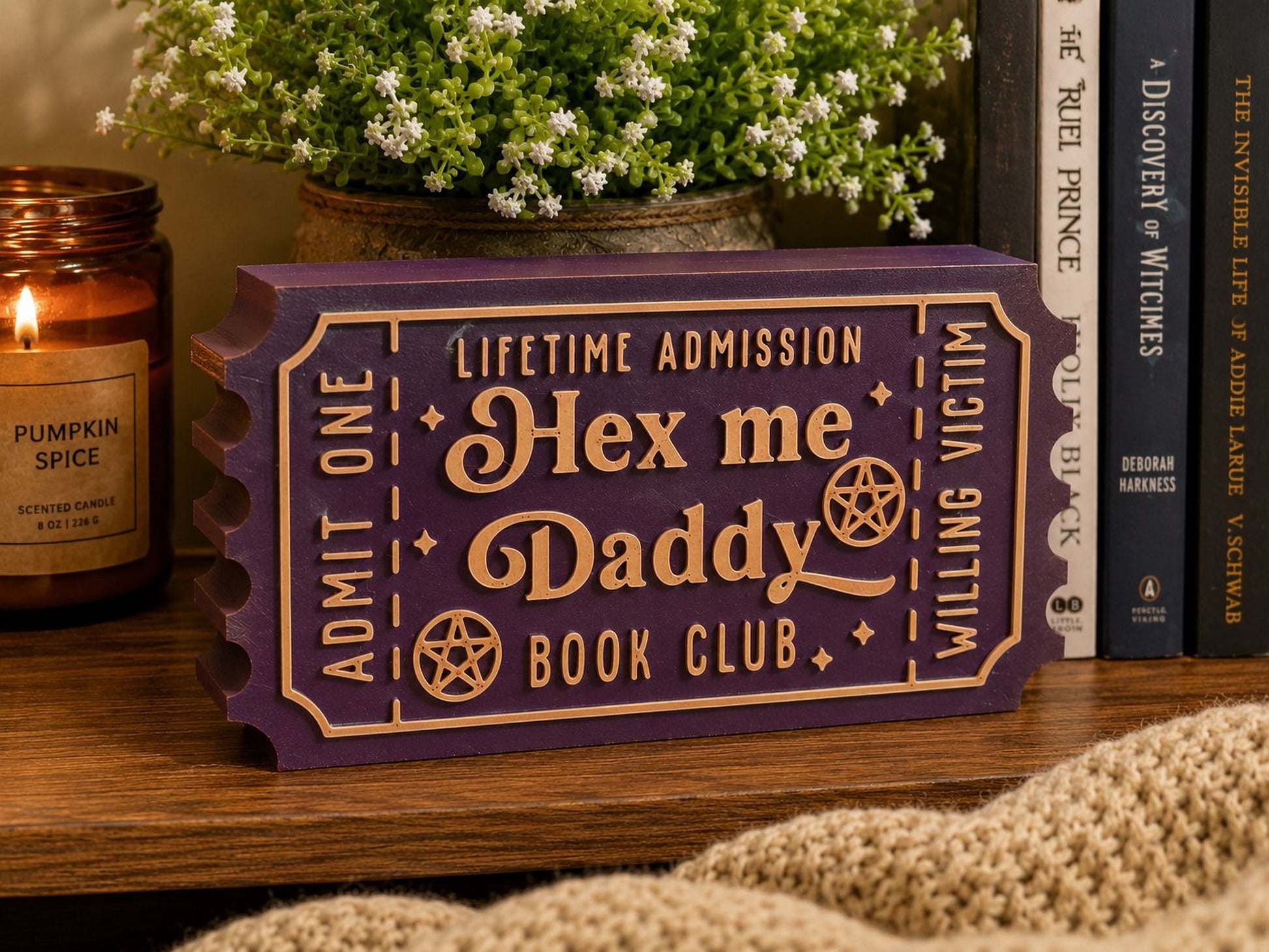 A purple ticket-shaped object with the text "Lifetime Admission Hex me Daddy Book Club" on it, placed on a wooden surface next to a candle and some books.