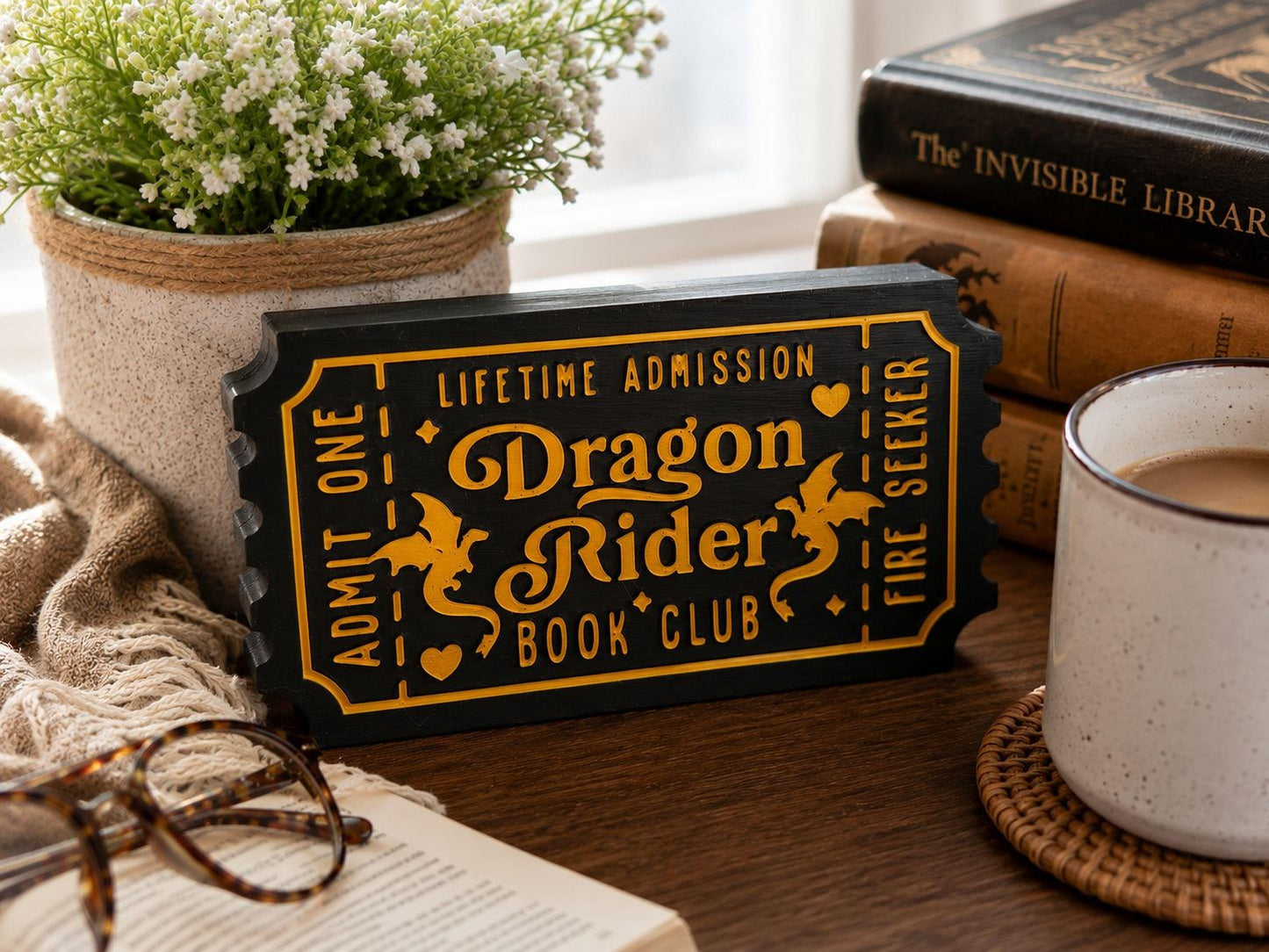 a black and gold ticket-shaped sign that reads "Dragon Rider Book Club" on a wooden surface, surrounded by a potted plant, a cup of coffee, and a stack of books.