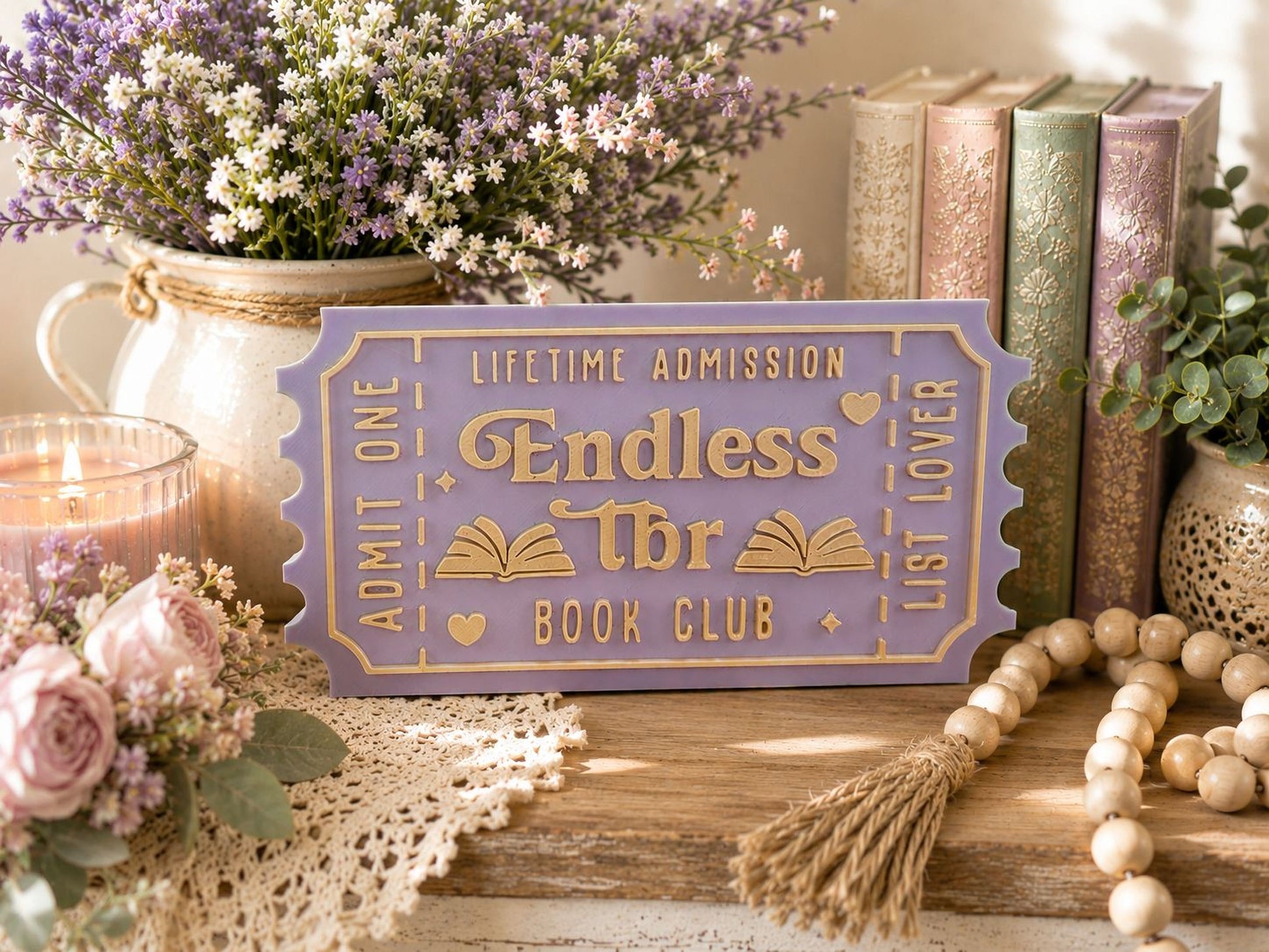 a purple ticket-shaped sign with gold lettering, surrounded by various decorative items such as books, flowers, and a beaded string.
