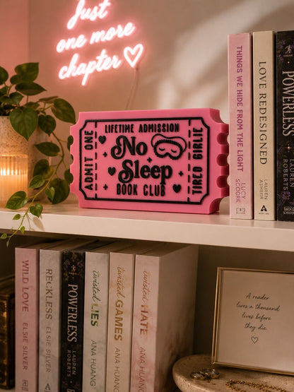 A pink ticket-shaped book club sign sits on a shelf among various books, with a neon sign reading "Just one more chapter" in the background.