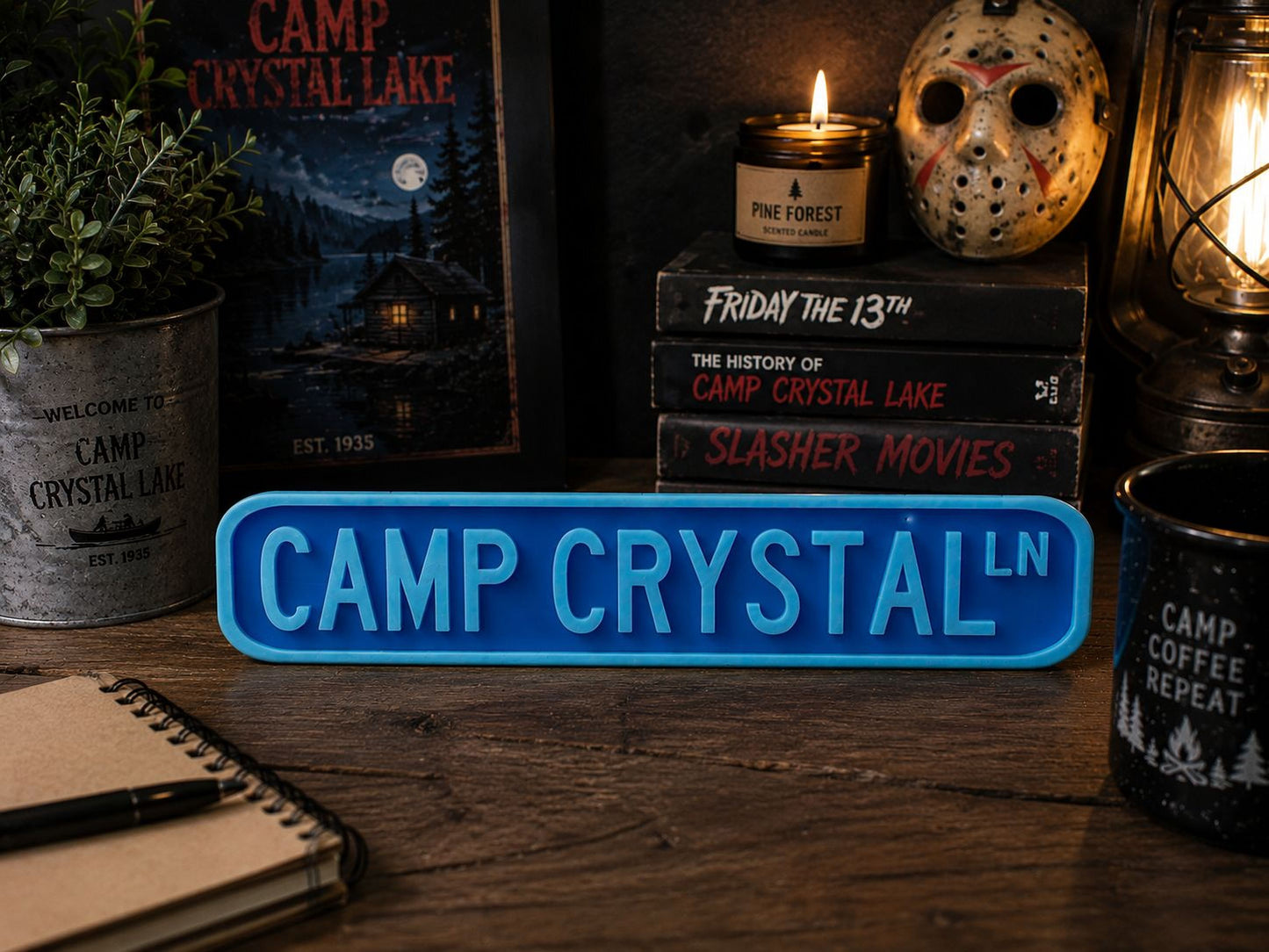 a collection of items related to the "Camp Crystal Lake" franchise, including a blue sign with the name "CAMP CRYSTAL", a stack of books, a candle, a mug, and a notebook.