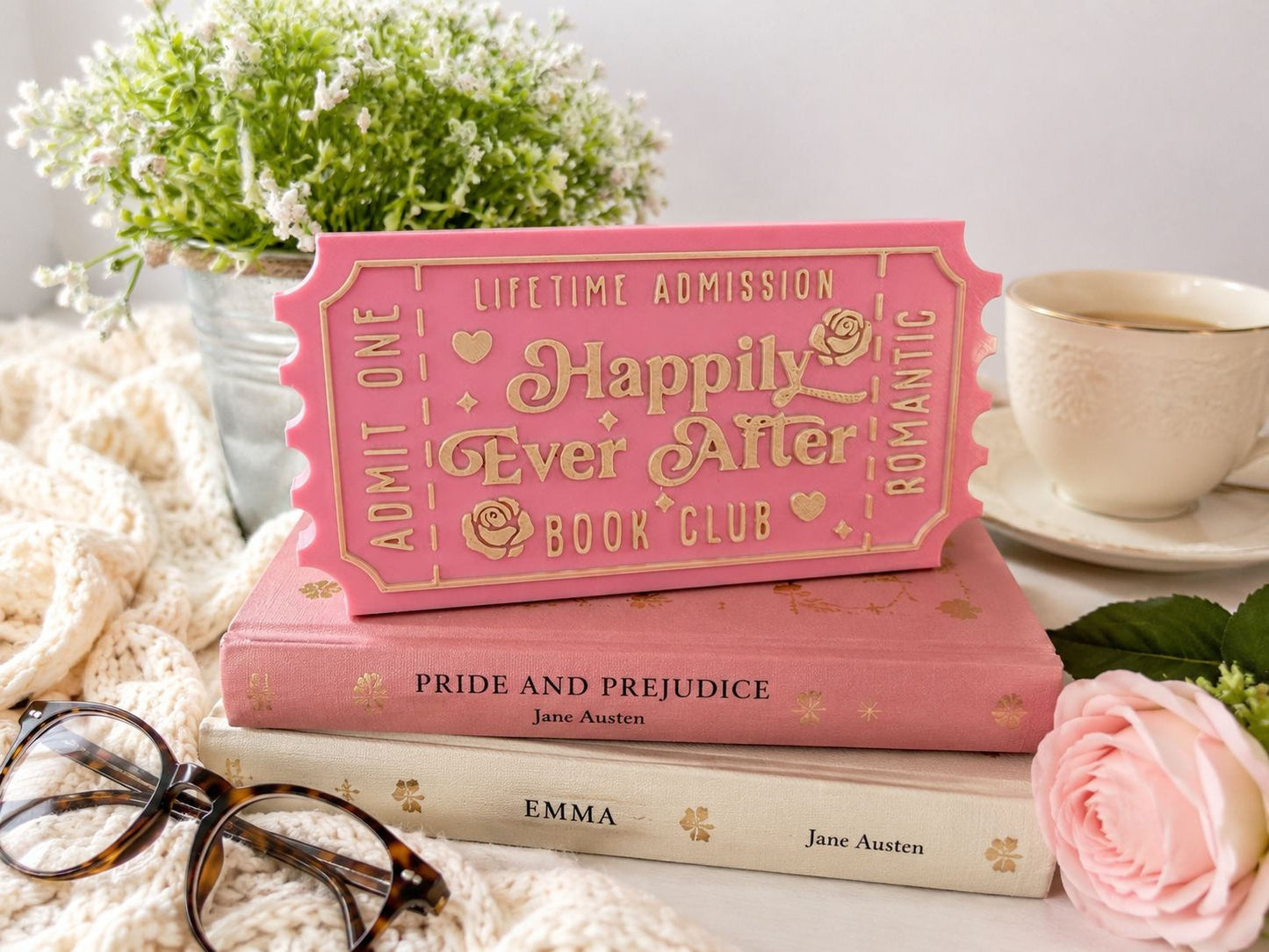 a pink ticket-shaped object with text, placed on top of two books, one of which is titled "Pride and Prejudice". The scene includes a cup of coffee, a rose, and glasses.