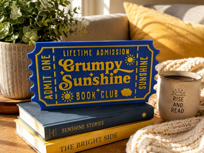 a blue ticket-shaped sign with the text "Grumpy Sunshine Book Club" on it, placed on top of a stack of books. There is also a mug with the text "RISE AND READ" on it, and a potted plant in the background.