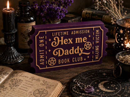 a purple ticket-shaped box with the text " Hex me Daddy Book Club" on it, surrounded by various items such as a candle, a book, and a crystal ball.