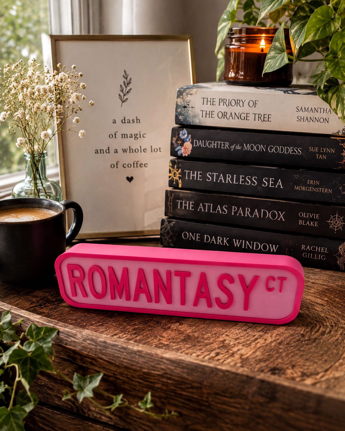 a wooden desk with a stack of books, a pink sign with the word "ROMANTASY" in large letters, a black mug, and a vase with white flowers.