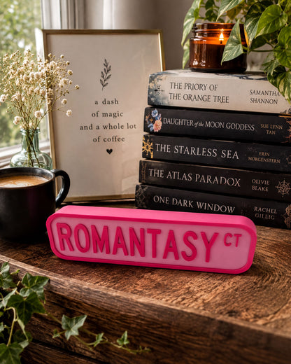 a wooden desk with a stack of books, a pink sign with the word "ROMANTASY" in large letters, a black mug, and a vase with white flowers.