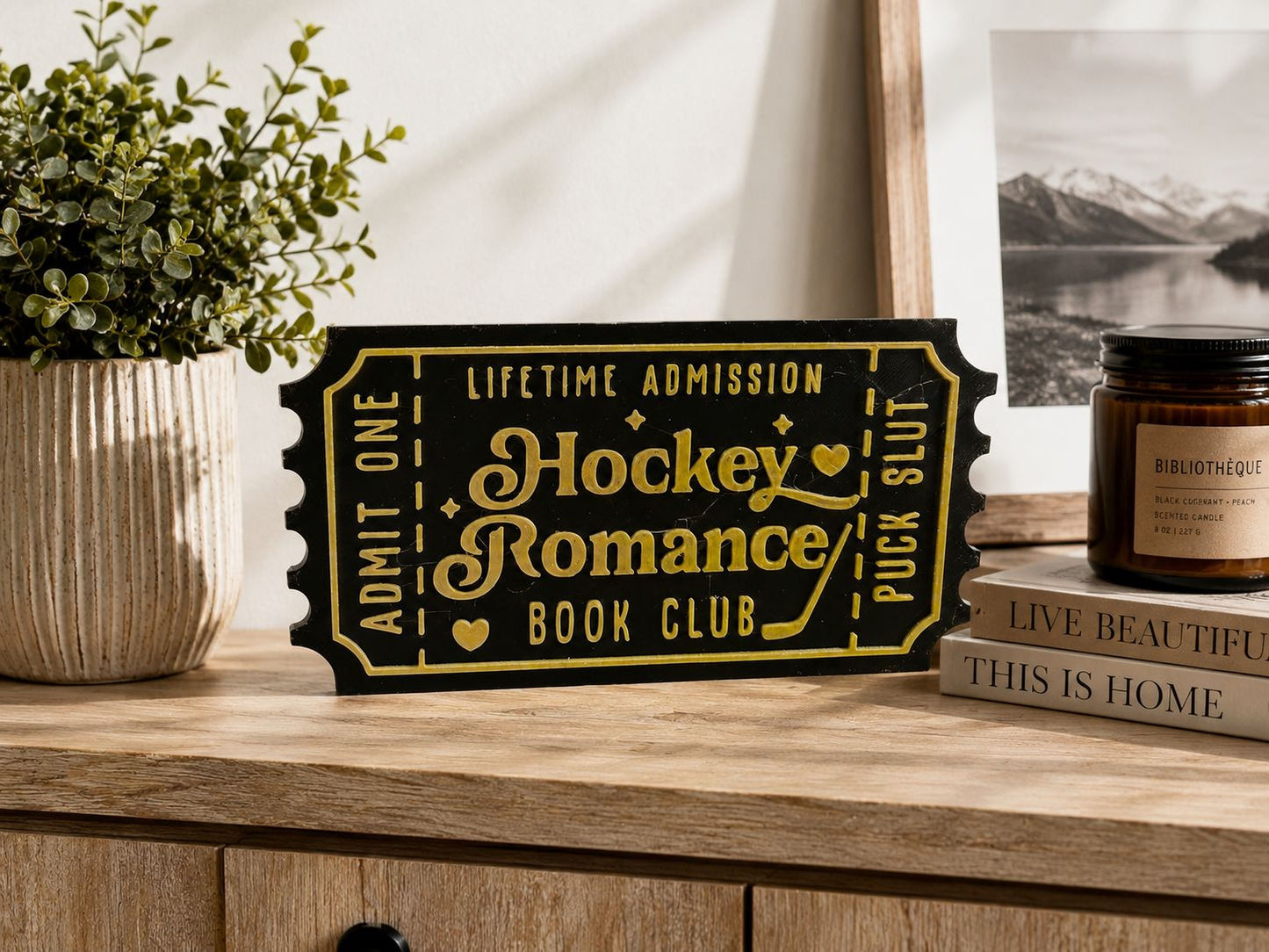 a wooden table with a black and gold ticket-shaped sign that reads "Hockey Romance Book Club" and a candle, along with a book and a plant.