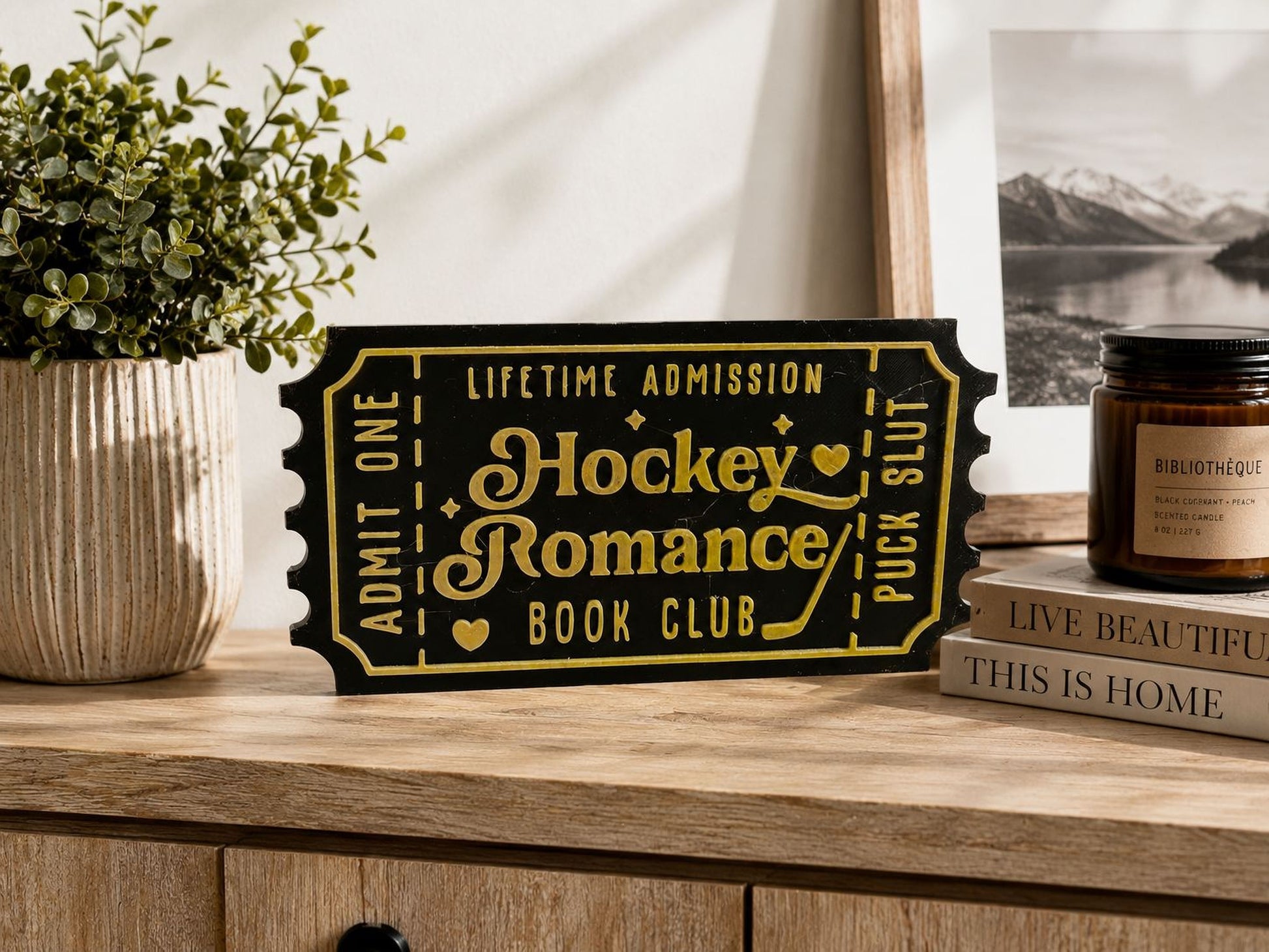 a wooden table with a black and gold ticket-shaped sign that reads "Hockey Romance Book Club" and a candle, along with a book and a plant.