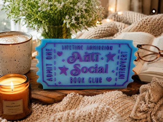 a cozy scene with a blue ticket-shaped object that says "Lifetime Admission Anti-Social Book Club", surrounded by various items such as a candle, a mug, a book, and a pair of glasses.