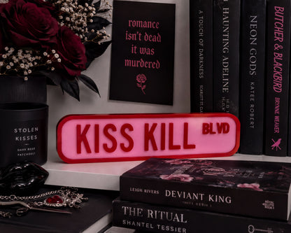 a collection of books, a pink sign, and some flowers arranged on a shelf.