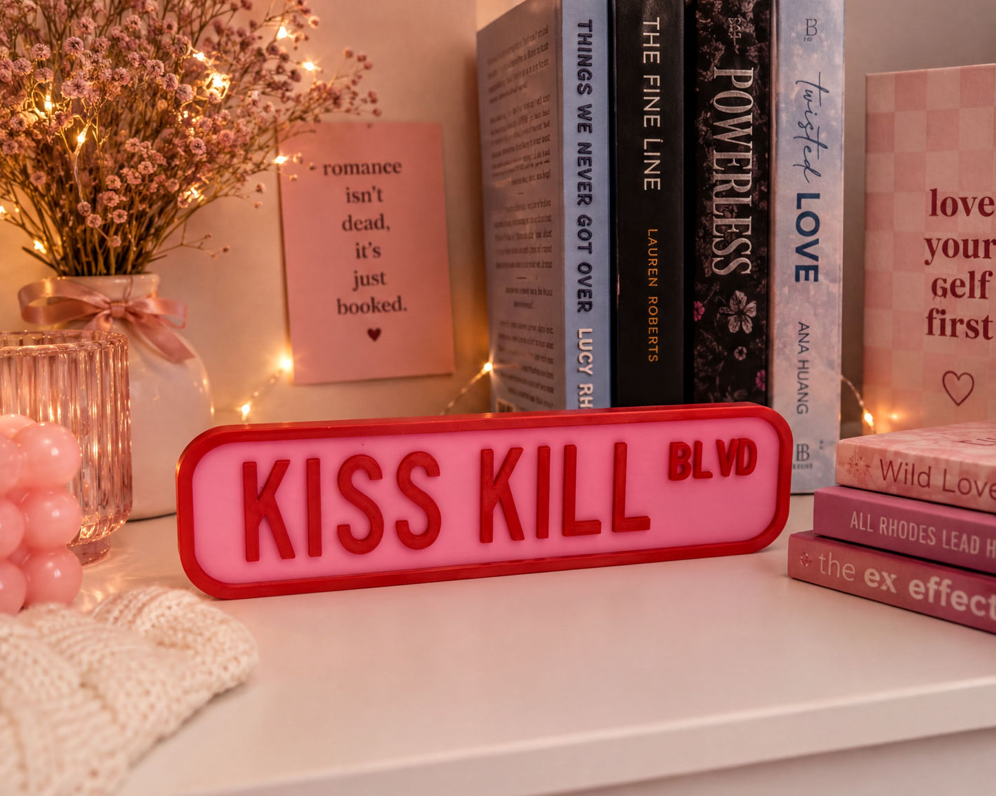 a pink sign with the words "KISS KILL BLVD" in bold red letters, placed on a white surface next to a stack of books. The books appear to be romance novels, and there is a vase of flowers and a string of lights in the background.