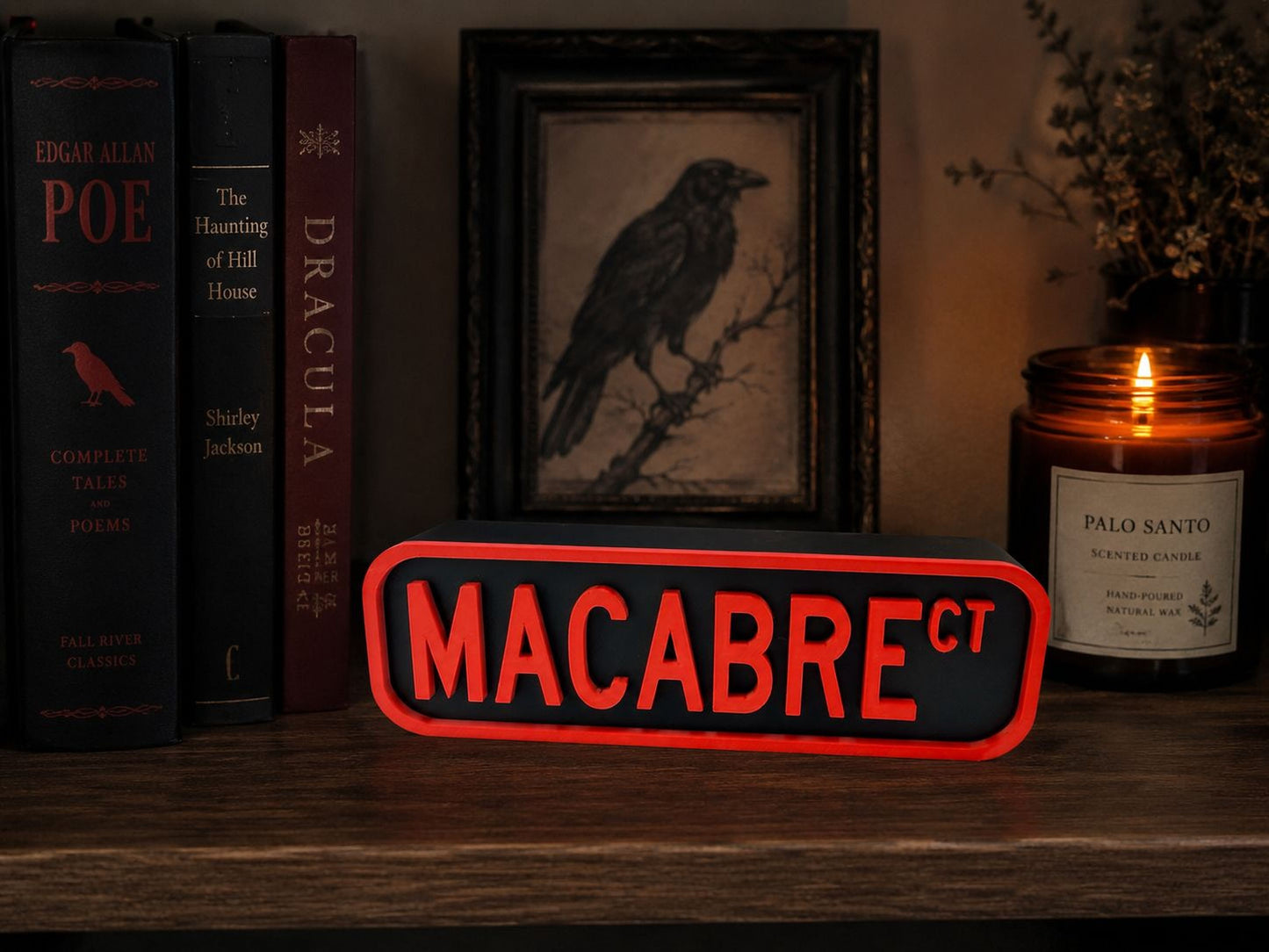 a bookshelf with various books, a framed illustration of a bird, a candle, and a red sign with the word "MACABRE" on it.