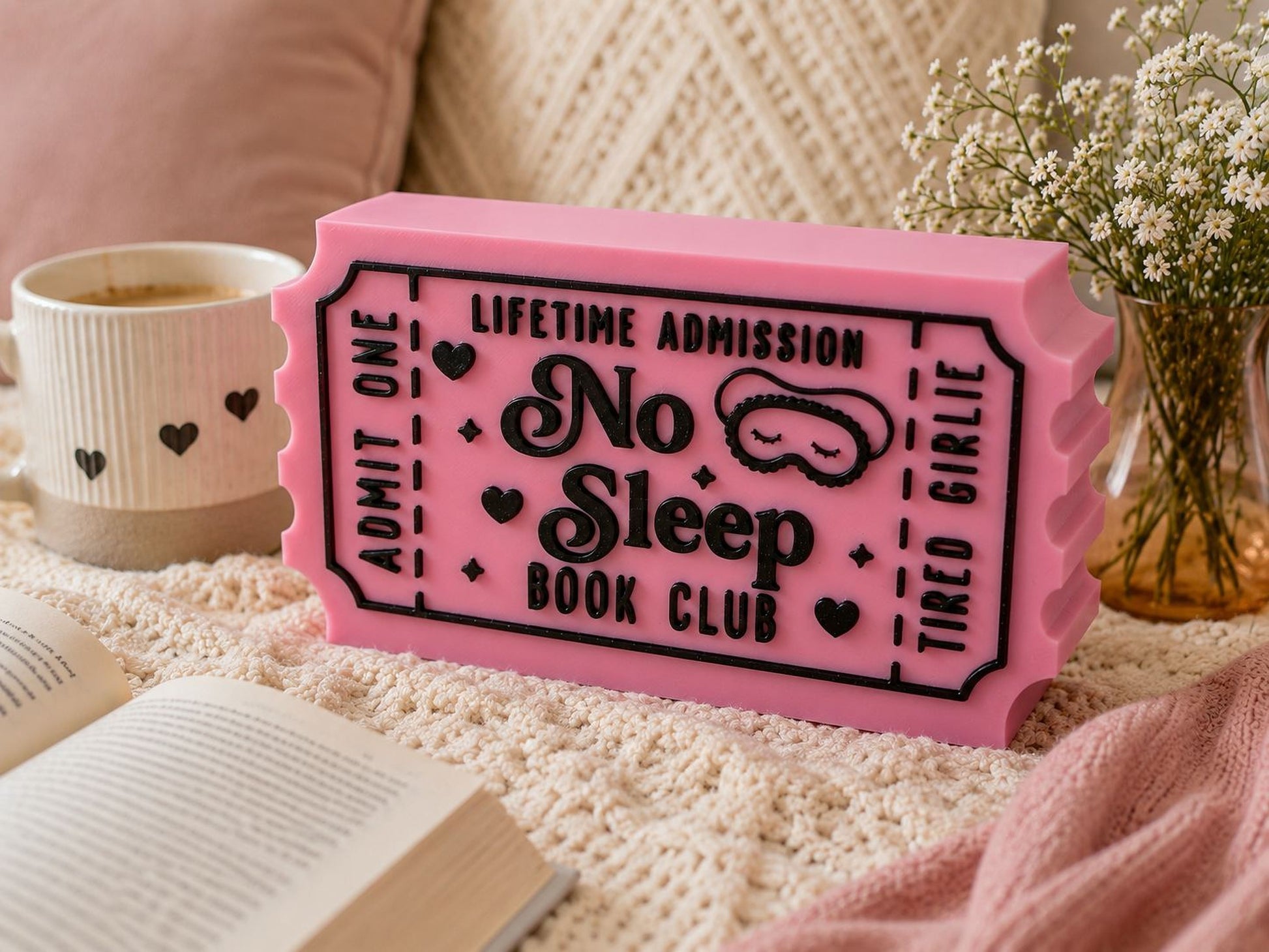 a pink ticket-shaped object with the text "No Sleep Book Club" on it, placed on a bed with a white blanket and a book.