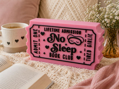 a pink ticket-shaped object with the text "No Sleep Book Club" on it, placed on a bed with a white blanket and a book.