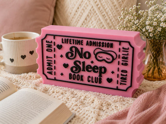 a pink ticket-shaped object with the text "No Sleep Book Club" on it, placed on a bed with a white blanket and a book.